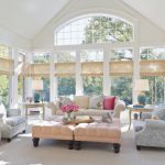 sunroom with white vaulted ceiling, windows around, blue sofas, soft orange ottoman for table, soft yellow sofa, pillows, rattan curtain, rattan round ottoman, side tables, table lamps