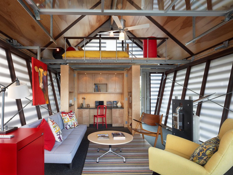 ecletic room in tiny house with yellw bed in battic bedroom with metal rail, red cabinet, grey cofa, stripped rug, metal beams, cream couch, wooden chair, wooden shelves