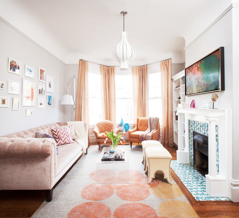 small living room with orange rug, soft orange gordyn, soft orange velvet sofa, orange chairs, white ottoman, clear acrylic coffee table, white shelves, firepla