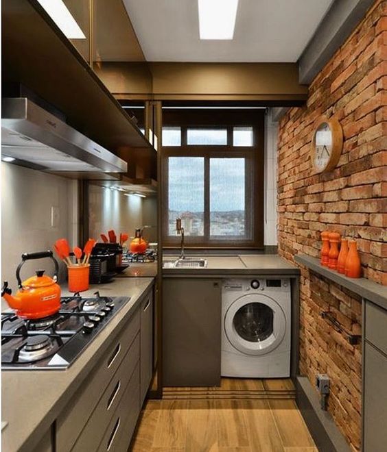 long kitchen with wooden floor, open brick wall, grey cabinet and top, washing machine under the top, near the sink