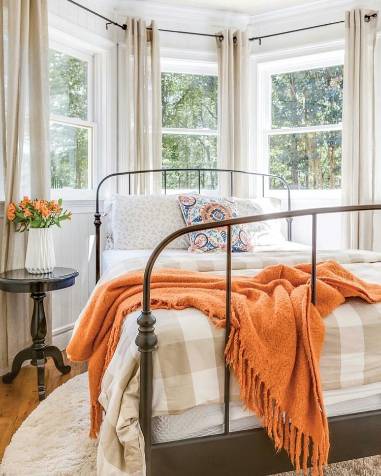 bedroom with wooden floor, white rug, metal bed platform with white bedding, pattern pillows, flowers, windows, curtains, orange blanket