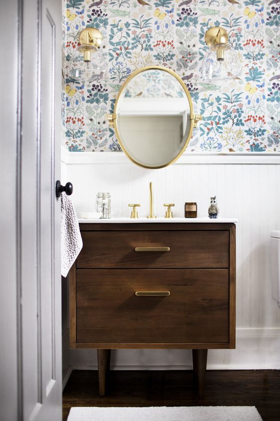bathroom, wooden floor, white wainscoting, flower pattern wallpaper, glass sconces, wooden vanity, golden framed mirror