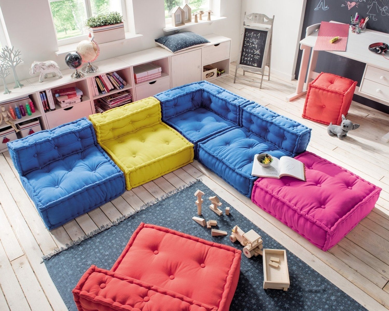 playroom, blue pink yellow floor sofa, wooden floor, white cabinet and shelves, white table, orange square ottoman, dark grey rug