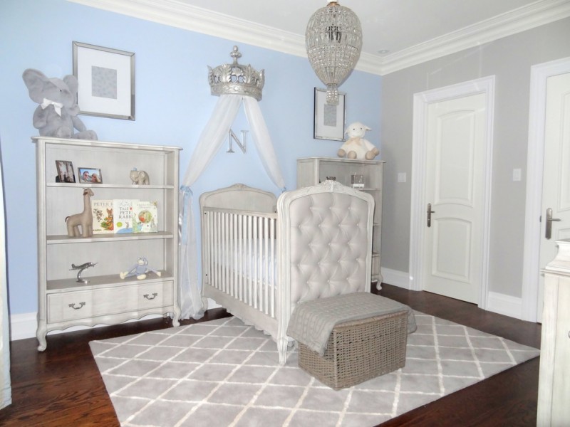 white theme room painted in light blue and grey with white cabinet for toys, white crib, white ottoman, silver king crown and curtain on top of the crib, crystal hanging lamp