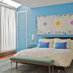bedroom with light blue wall, wooden flooring, wooden storage chest with blue cushion, white curtain, white framed blue door, white bed cover with white and blue pillows, grey rug