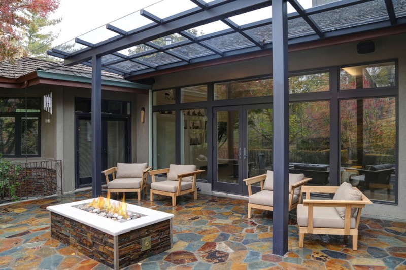 expensive and luxurious backyard patio with dark toned pergola covered with transparent glass panels wooden chairs with white puff comforters center fire pit table slate floors