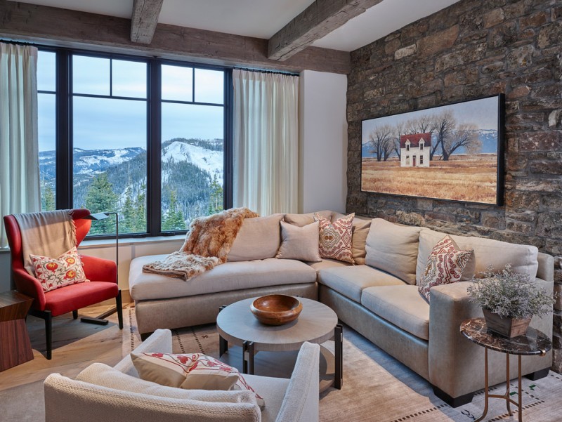 mountain style living room idea neutral toned sectional red corner chair neutral toned rug neutral toned table in round shape hard textured stone walls white ceilings with exposed logs