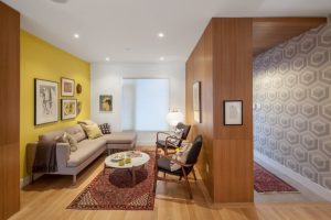 small livingroom with wooden wall, yellow wall, white wall with distorted glass window, beige corner sofa, white round coffee table, red rug, black chairs