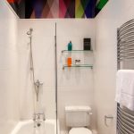 white small bathroom with colorful assymetric pattern ceiling, white toilet, white sink, white tub with showe and glass partition, glass shelves