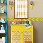 Bathroom With Beige Color, Blue Shelves, Yellow Lining On The Wall, Mirror, Yellow Cabinet With Ikat Pattern On The Door, Yellow Sconces