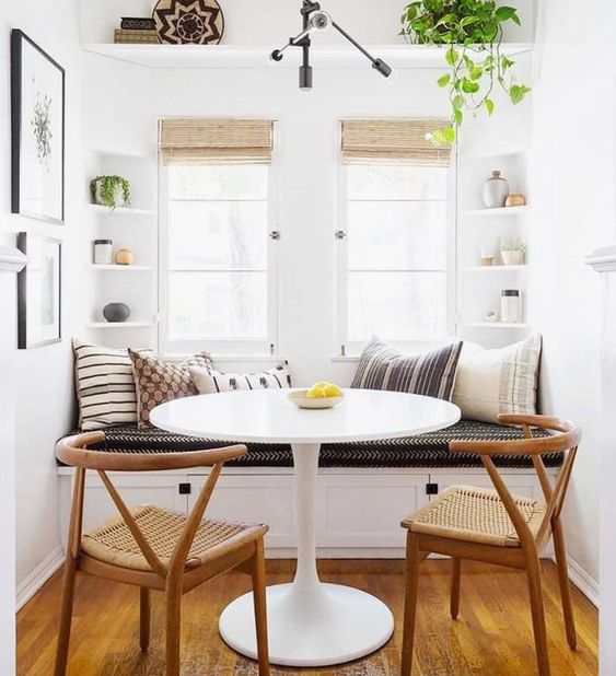 corner dining nook with white bench with cushions and pillows, white round table, brown rattan chairs, built in small corner shelves on two corner, windows