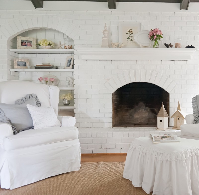 paint fireplace white brick wooden floor rattan rug white armchair white skirt lounge chair white built in shelves wood beams pillows