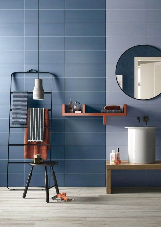 blue wall tiles, light blue backsplash wall, round mirror, wooden bench, white round tall small sink, black rack, black stool, wooden floor