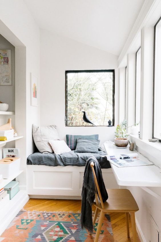 window nook, white built in bench, grey cushion, white floating table, wooden chair, wooden floor, colorful rug, white wall,