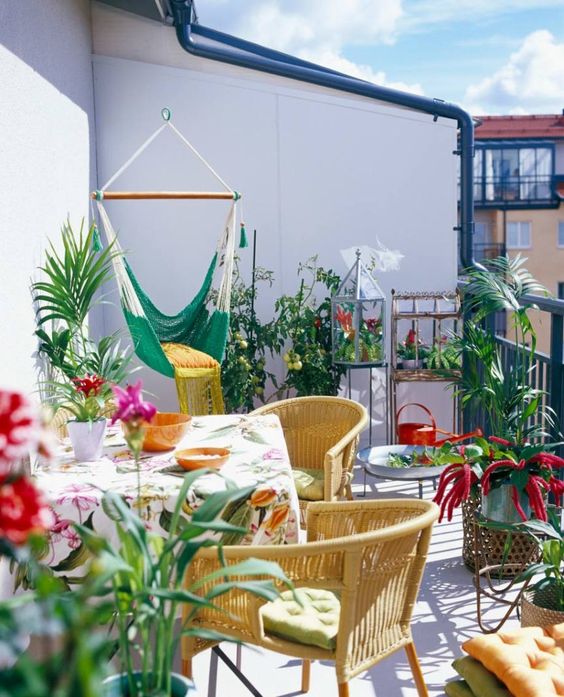 balcony, white floor, white wall, green swing, rattan chairs, table with colorful clothe, plants, rattan pots, pillows