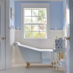 Bathroom, White Wainscoting, Blue Wall, White Framed Window, White Tub With Golden Claw Foot, White Wooden Floor, Blue Patterned Rug, White Side Table