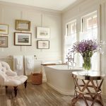 Bathroom, Wooden Floor, White Wainscoting, White Tub, Golden Side Table, White Tufted Chair, White Shade On The Window, Paintings, Rattan Basket